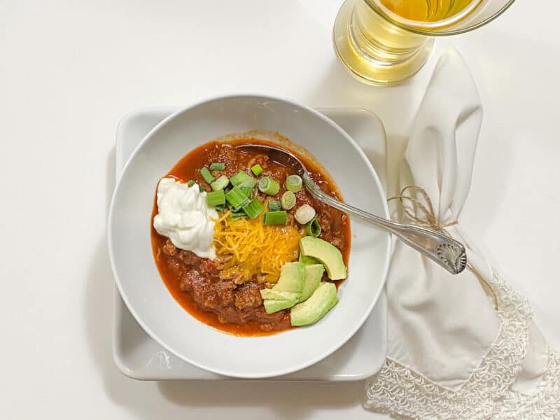 White bowl on a white plate filled with chili . White napkin on the right side with a large soup spoon and a beer glass with a cold beer. All ready for the perfect meal for a cold winter day or a football game