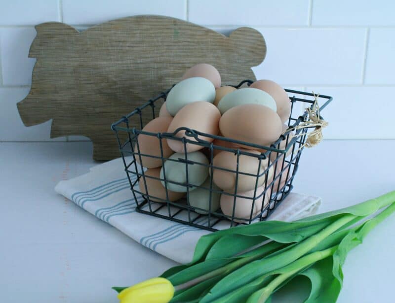 picture of fresh eggs in a wire basket on the counter with a pig cutting board, a blue and white striped towel and yellow tulips