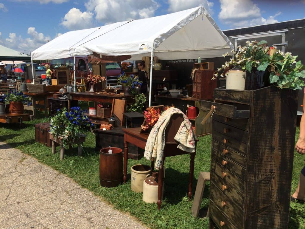 Outdoor flea market booth under a white tent displaying vintage furniture and cabinets—perfect for creating a pantry in unused space—with decorative items, potted flowers, and ceramic jugs on a grassy area surrounded by people and tents.