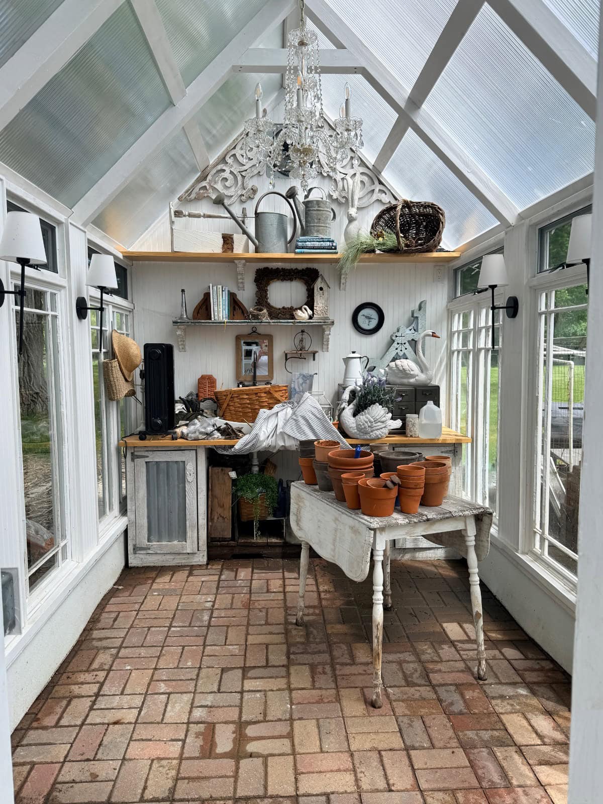 A bright, airy greenhouse with a brick floor, glass roof, and white walls. A rustic table holds terra cotta pots and gardening tools; shelves at the back display baskets, books like And The River Rises, and decor. A chandelier hangs from the ceiling.