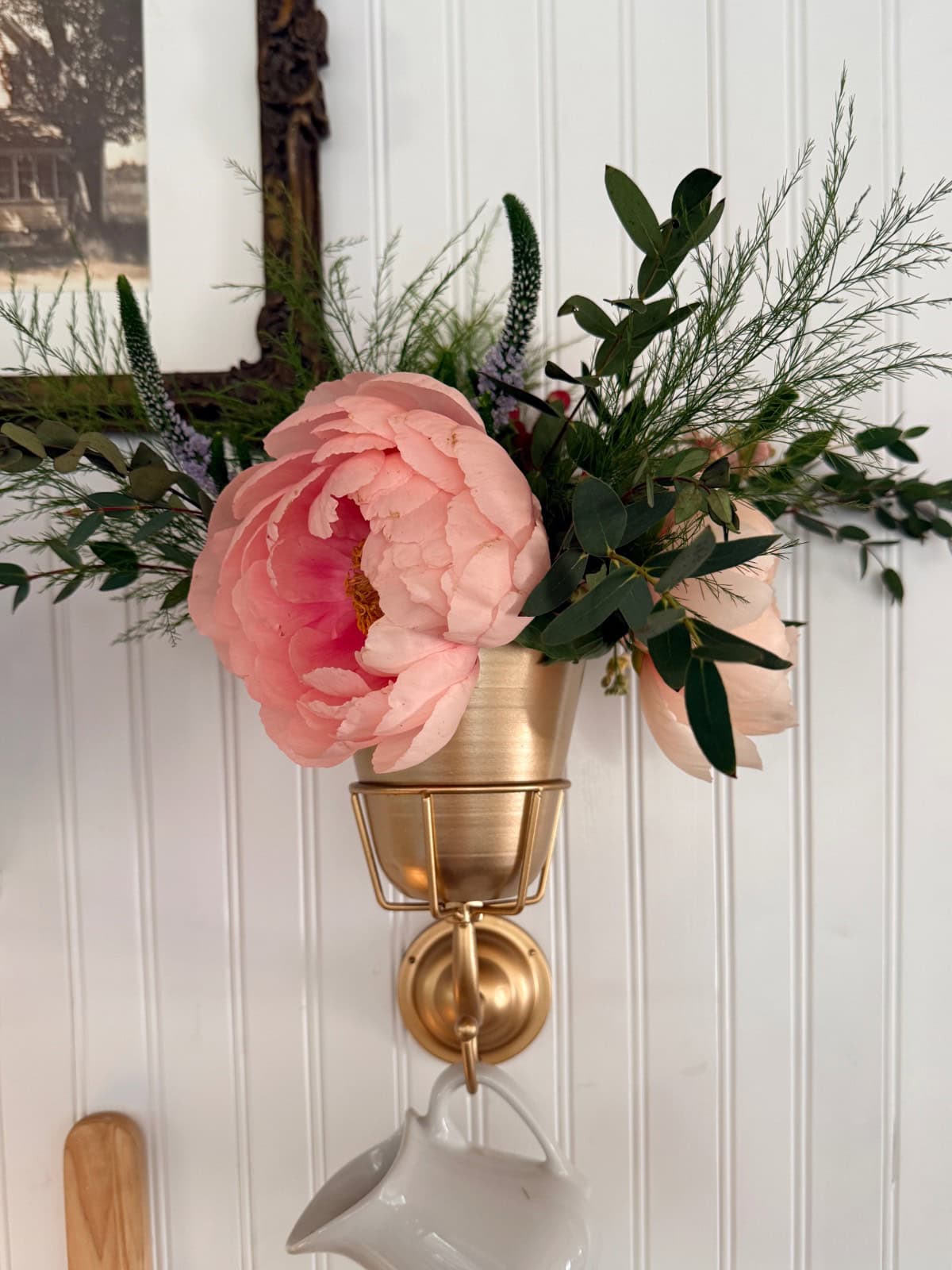 A gold wall vase holds pink peonies and assorted greenery against a white beadboard wall, with a white pitcher hanging from a hook below. The scene evokes tranquility—like the gentle calm before And The River Rises in the framed picture nearby.