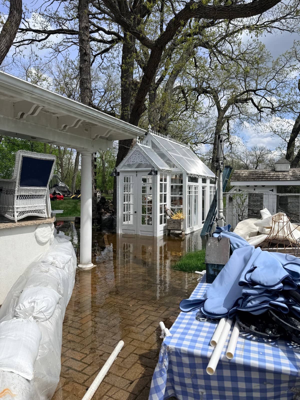 A flooded backyard with water covering the patio, as And The River Rises. A small white greenhouse stands among tall trees. Outdoor furniture, sandbags, and blue-and-white checked tablecloths are seen in the foreground.