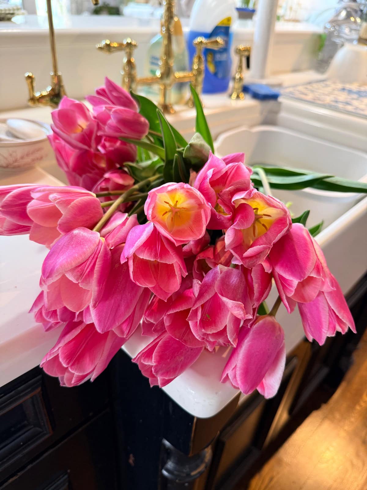 A bouquet of vibrant pink tulips with yellow centers rests on the edge of a white kitchen sink, gold faucets gleaming nearby—an inviting scene where beauty blooms, as if And The River Rises just outside the window.