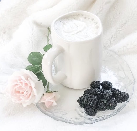 A white mug of frothy milk sits on a clear glass plate with fresh blackberries and a pale pink rose, all set on a soft white fabric background. What are these empty nesters planning to do as they enjoy this peaceful morning?.