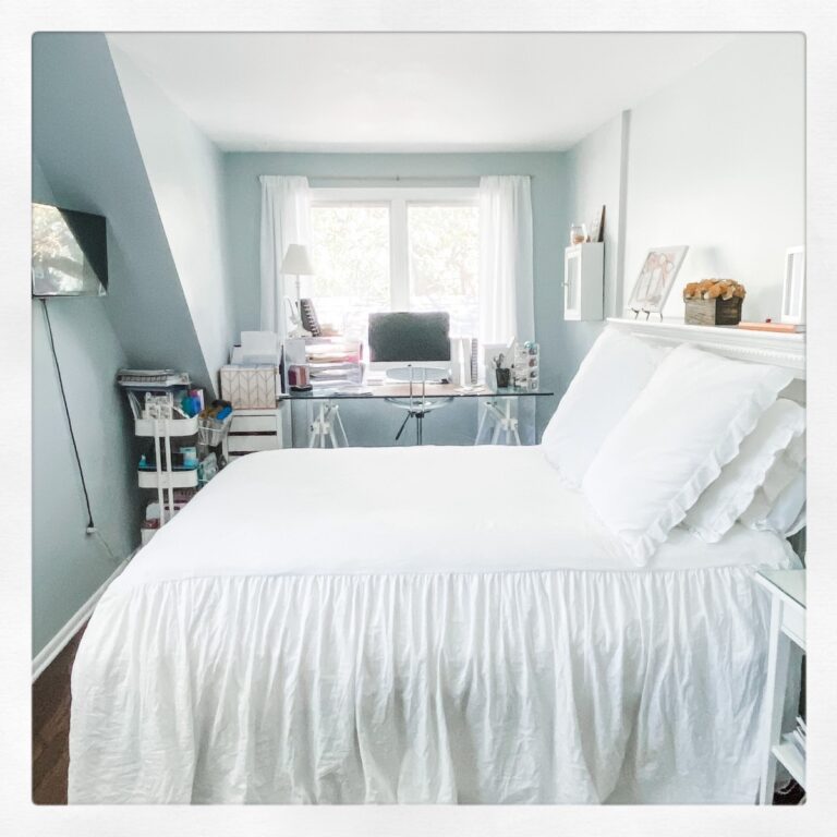 A bright bedroom with a neatly made bed in white linens, a desk under a sunlit window, and organized shelves—what are these empty nesters planning to do in such a cozy, airy atmosphere.