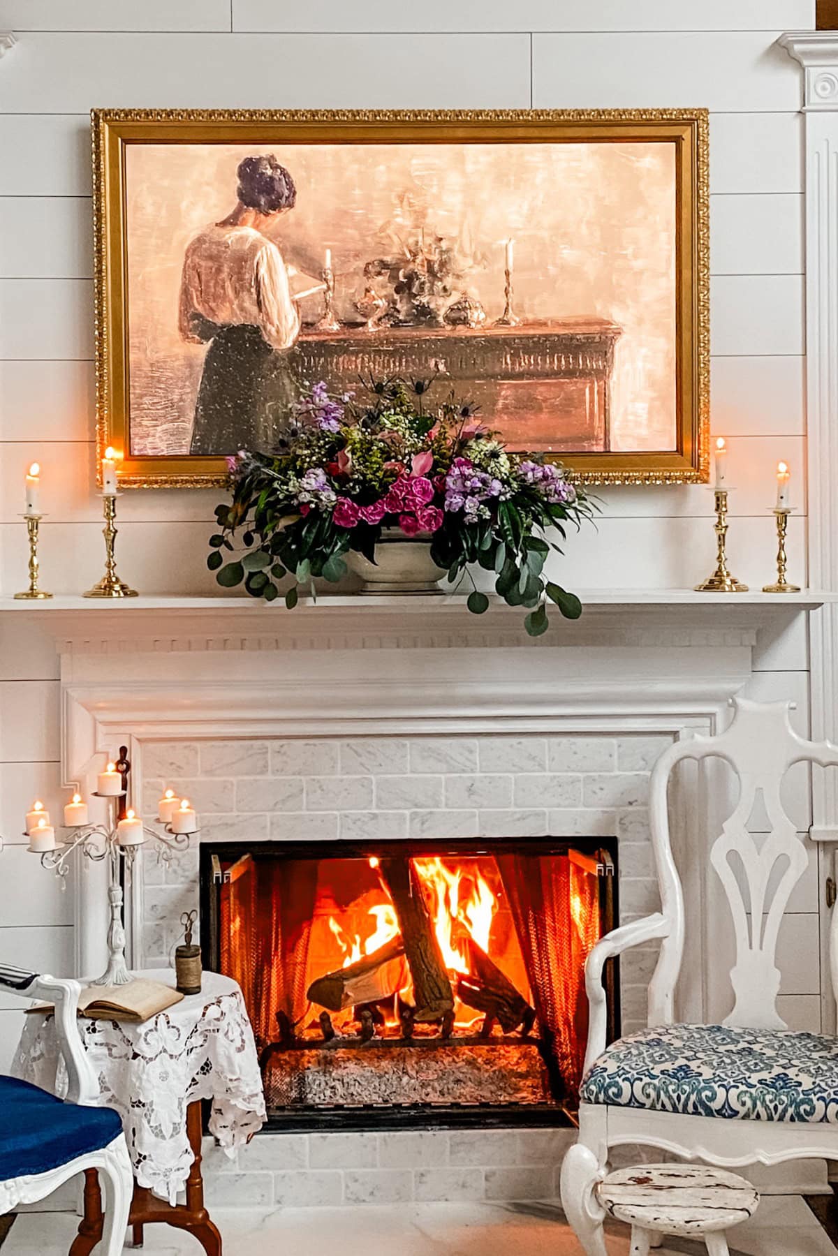 A cozy fireplace with a roaring fire, flanked by white chairs. Above the mantel are gold candlesticks, a lush floral arrangement, and a framed painting—perfect for inspiring your Fall House Tour 2017 décor.