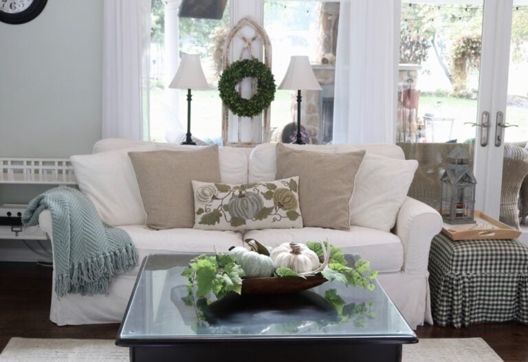 A cozy living room with a white sofa, beige and green pillows, a light blue throw, and a decorative pumpkin pillow. As part of the Fall House Tour 2017, pumpkins and greenery on the glass coffee table add seasonal charm.