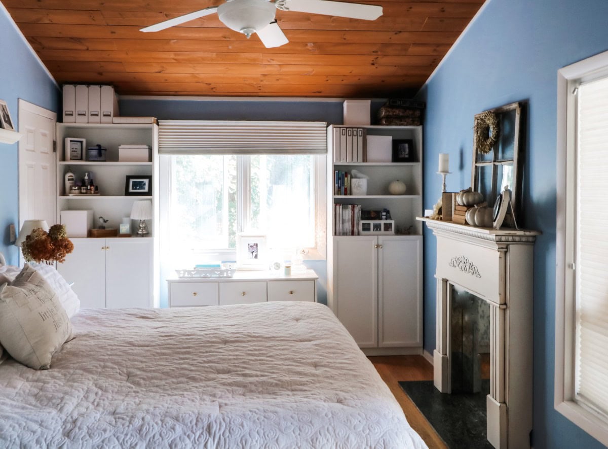 A cozy bedroom from the Fall House Tour 2017 features blue walls, a wooden ceiling with a white ceiling fan, a bed with a white quilt, built-in shelves and cabinets, a fireplace, and a large window letting in natural light.