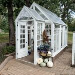A white greenhouse with glass walls, pumpkins, and potted plants on a brick patio—autumn is the season here.