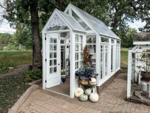 A white greenhouse with glass walls, pumpkins, and potted plants on a brick patio—autumn is the season here.