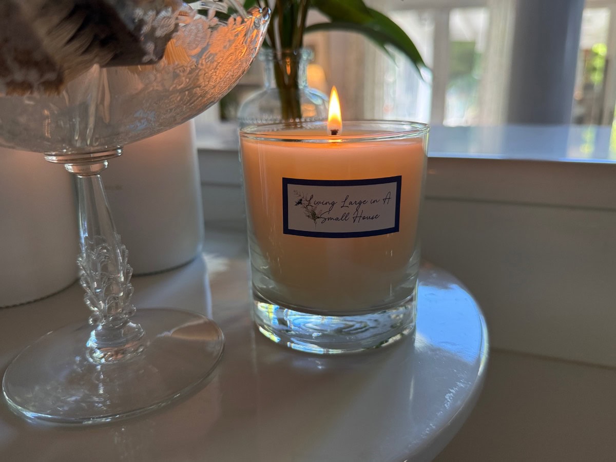 Lit candle in a glass jar labeled “Always Leave a Small House” evokes cozy Autumn is the Season vibes on a white table.