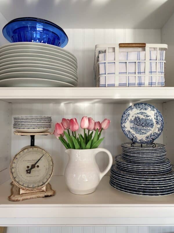 open shelves with dishes stacked, basket with towels, vintage scale and an ironstone pitcher with pink tulips in it.