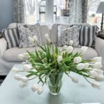 A glass vase of white tulips sits on a coffee table in front of a white sofa with patterned pillows, adding a homemaker’s touch to a cozy, well-lit living room with blue accents and large windows in the background.