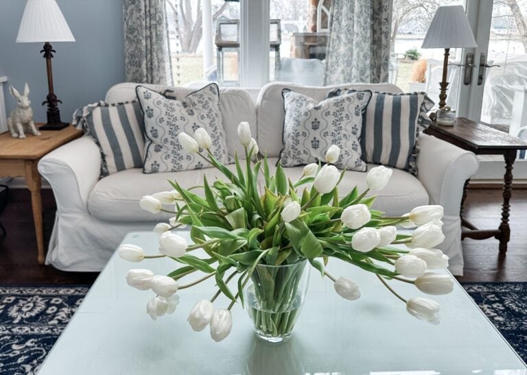 A glass vase of white tulips sits on a coffee table in front of a white sofa with patterned pillows, adding a homemaker’s touch to a cozy, well-lit living room with blue accents and large windows in the background.