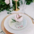 wedding table setting with rose gold chargers, topped white dinner plate, vintage pastel floral salad plate and vintage teacup that has a peony and wooden laser name tag in the cup.