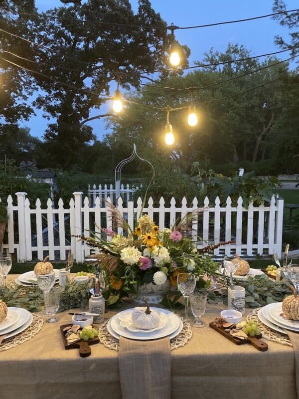 Inspired by How to Dine Al Fresco with Fall Decor, an outdoor dining table is decorated with a beige tablecloth, white plates, pumpkin-shaped napkins, flowers, and greenery. String lights twinkle above near a white picket fence.