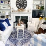 Farmhouse living room with white and navy rug. White sofa and chair and a blue and white checked chair surrounding white fireplace flanked by white shelves