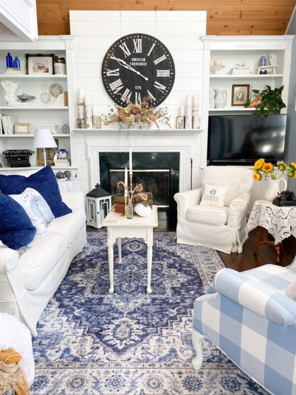 navy and white area rug in a cozy farmhouse living room with a white sofa and chair. There is also and a white and blue checked chair. The furniture surrounds a white fireplace with a black farmhouse clock hanging above the mantle