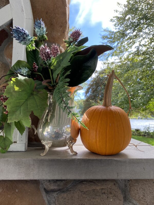 silver pitcher filled with faux fall foliage and a pumpkin from my garden on our outdoor fireplace mantel. Beautiful blue sky and blue water in the background