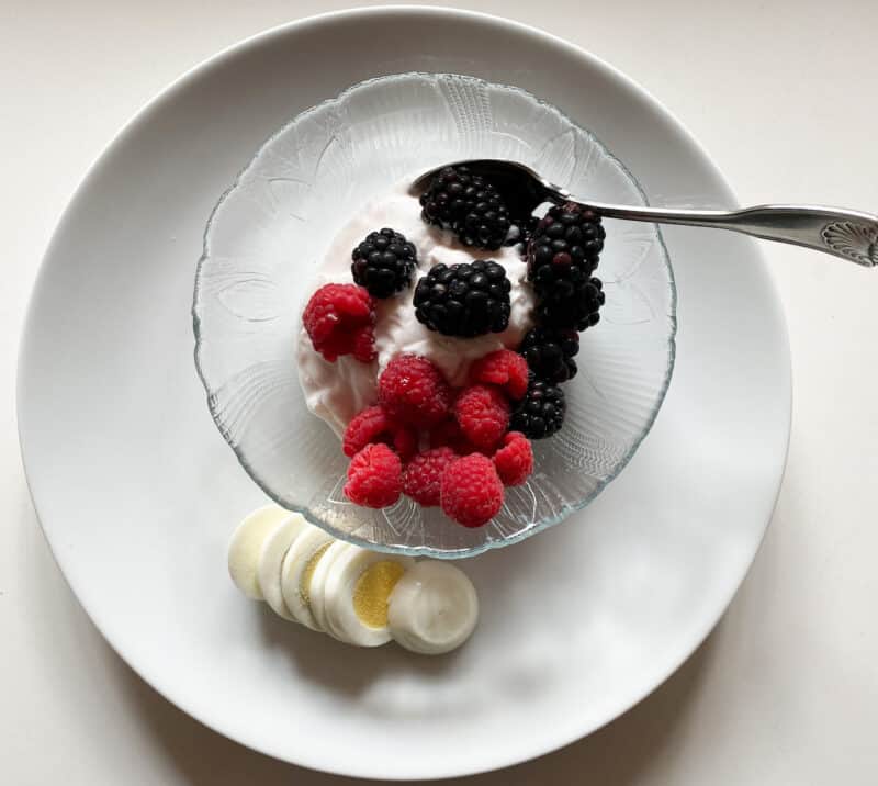 Yogurt with raspberries and blackberries in a glass bowl on a white plate with a hardboiled egg on the plate
