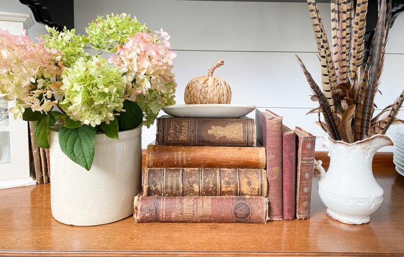 vignette with a crock filled with hydrangeas, vintage books topped with an ironstone dish along side a vintage either that hold pheasant feathers