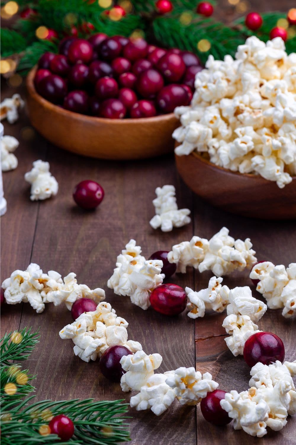 Bowls of fresh cranberries and popcorn sit on a wooden surface, with scattered popcorn, cranberries, and evergreen branches nearby—perfect for anyone wondering how to have a quiet little Christmas with cozy, festive touches.