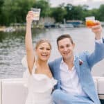picture of a bride and groom in the back of a boat on a river with their cocktails in the air - cheers