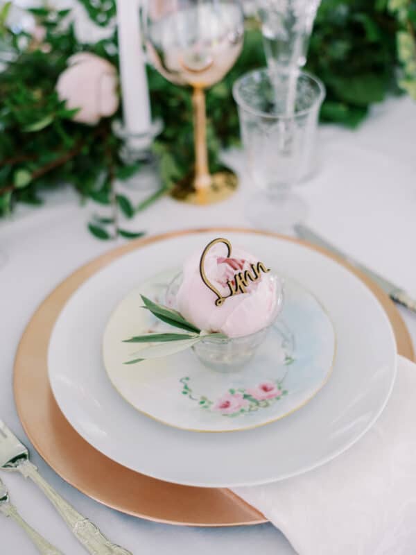 wedding table setting with rose gold chargers, topped white dinner plate, vintage pastel floral salad plate  and vintage teacup that has a peony and wooden laser name tag in the cup.