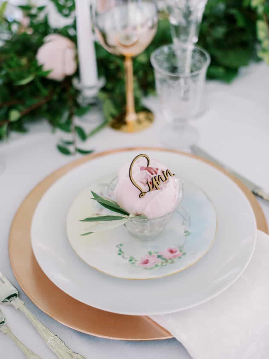 wedding table setting with rose gold chargers, topped white dinner plate, vintage pastel floral salad plate and vintage teacup that has a peony and wooden laser name tag in the cup.