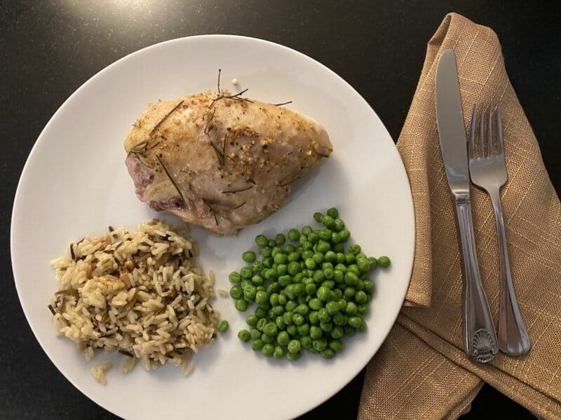 White plate on black counter with fork and knife on burlap napkin. On the plate is herb roasted bone-in chicken breast, wild rice and peas.