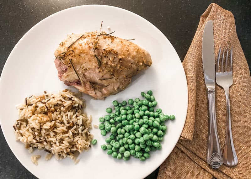 White plate on black counter with fork and knife on burlap napkin. On the plate is herb roasted bone-in chicken breast, wild rice and peas.