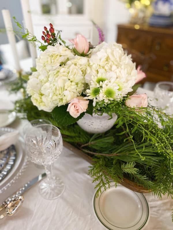 Our Christmas Eve Dinner Table with a dough bowl filled with winter greens and a cement vase inside with white hydrangeas and pink roses