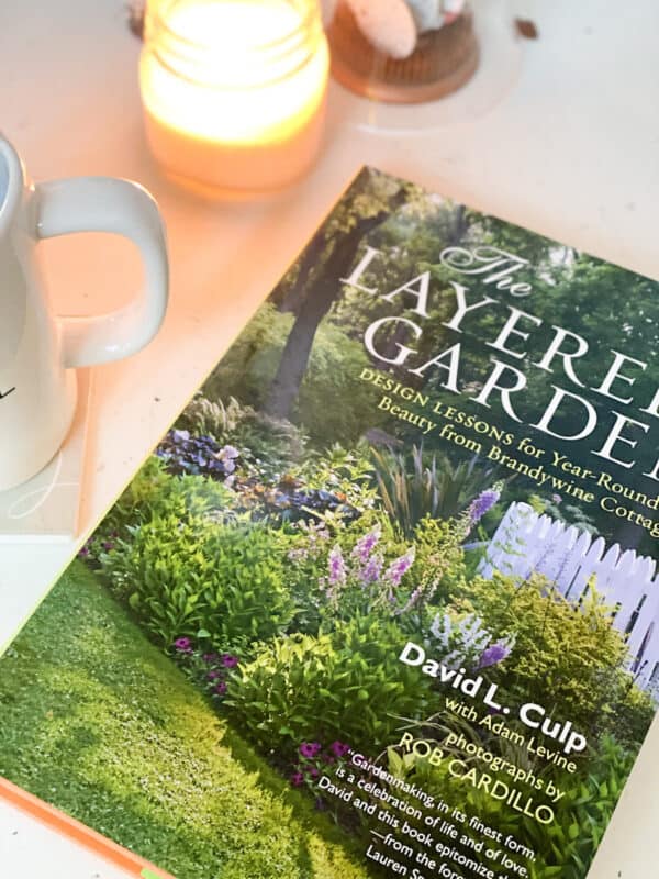 the book the layered garden on a table with a cup of coffee and a candle