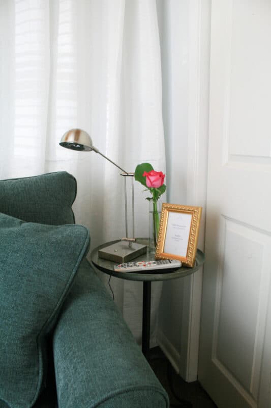 Small table next to sofa in guest room with a light, flower, wifi password in frame and tv remote control