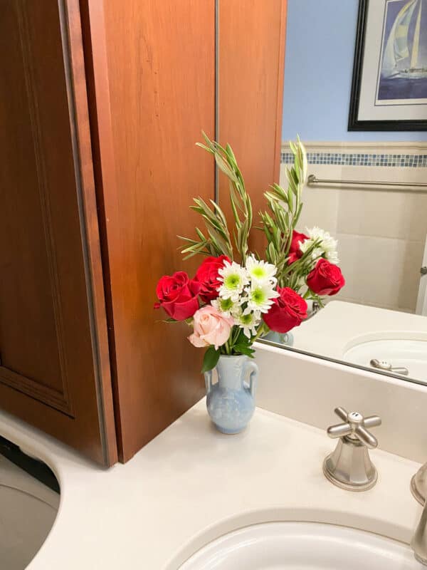 photo of the corner of the bathroom sink and clean mirror with a small blue bud vase in the corner. The vase has red and pink roses, white daisies and olive branches in it.
