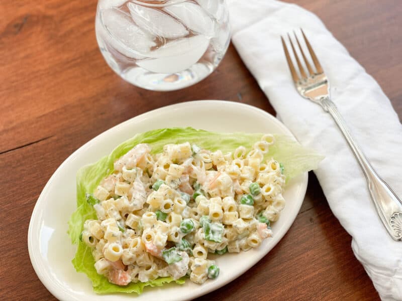 pasta and shrimp salad on top of a piece of lettuce on an oval plate. There is a white napkin with a fork on it to the right of the plate and a clear glass stemless glass with water and ice right above.