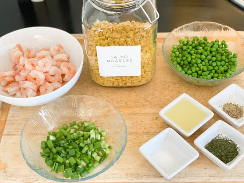 Cutting board with the ingredients for Pasta and Shrimp Salad layed out. Ther are Salad Noodles, Peas, Green onions, shrimp, salt, pepper, dill and lemon juice all in glass bowls or dishes.