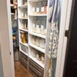Organized pantry under stairs with labeled glass jars, wooden crates below, and hanging aprons and tote bags on the right wall.