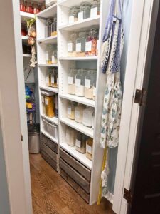 Organized pantry under stairs with labeled glass jars, wooden crates below, and hanging aprons and tote bags on the right wall.