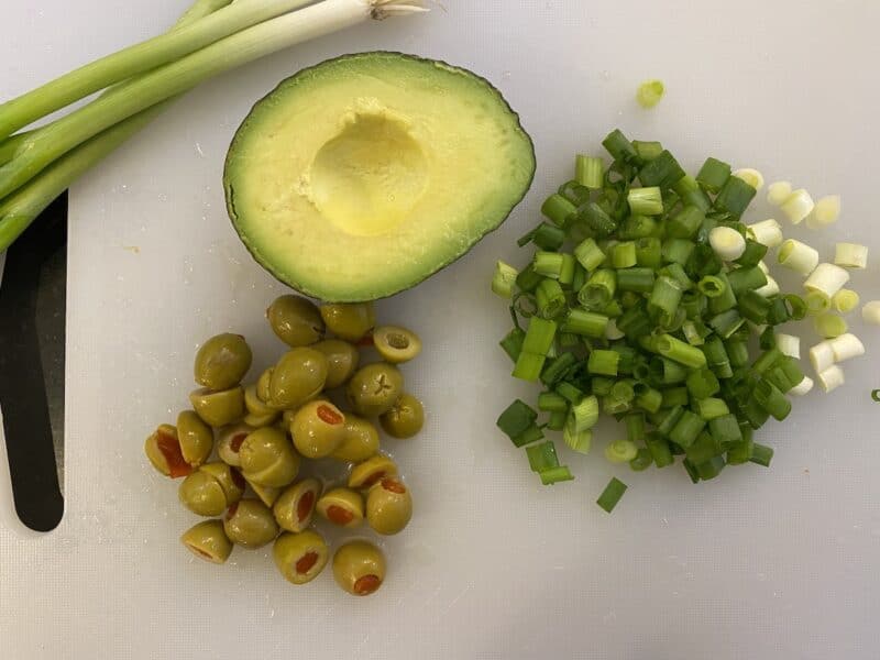 cutting board with chopped green onion, chopped green olives, half of an avocado and some whole green onions on the side