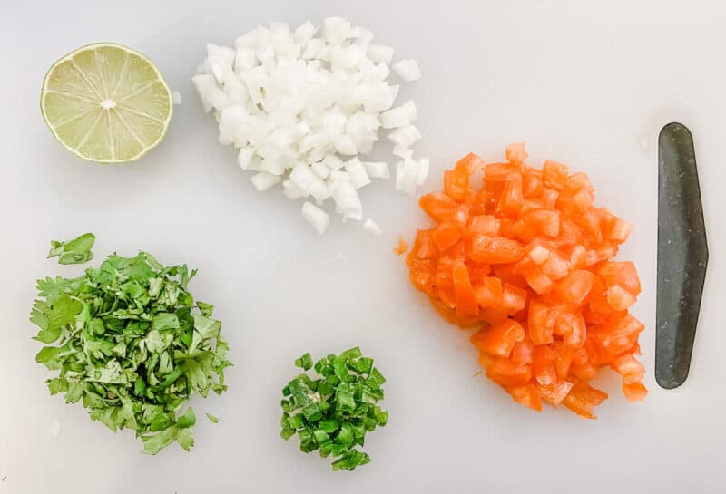 cutting board with all of the ingredients for pico de Gallo. Chopped tomatoes, chopped onion, chopped jalepanos, chopped cilantro and 1/2 of a lime
