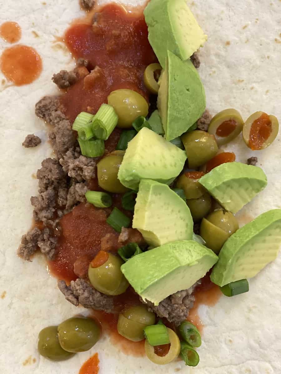 Unique Taco with a tortilla shell topped with browned ground beef, tomato sauce, chopped green onions and olive, and diced avocados