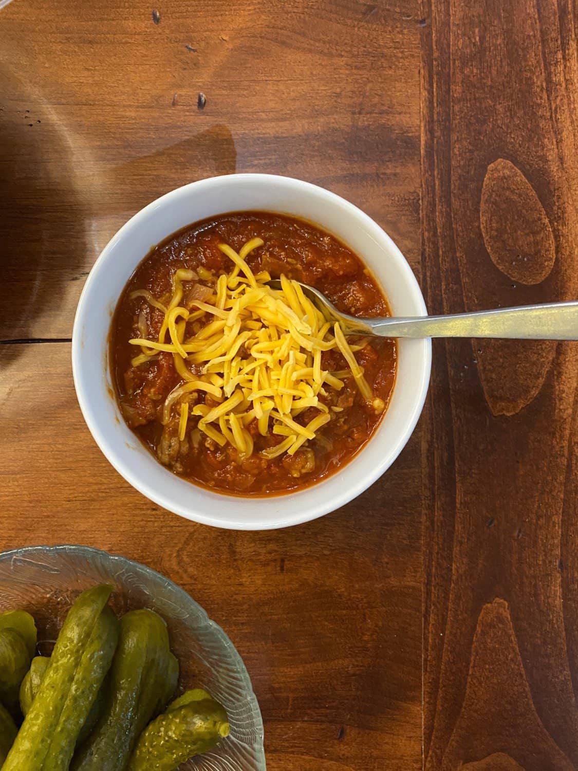 A bowl of turkey chili topped with shredded cheese and a spoon rests on a wooden table. To the side, there's a glass dish with pickles.