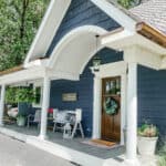 A charming house with dark blue siding and white trim, perfect for easy home renovation management. The front porch features a wooden door with a wreath, two striped chairs, potted plants, and a "Welcome" sign. A lantern hangs from the arched roof, framed by trees in the background.