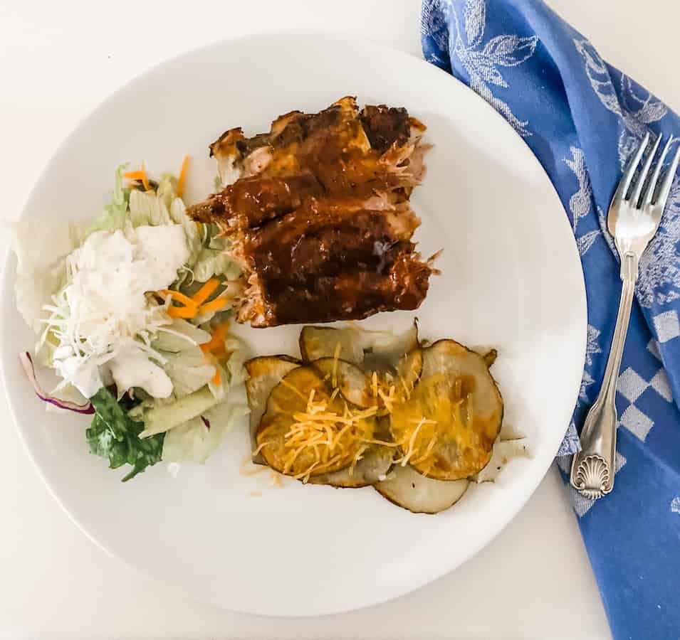The easy melt in your mouth rib recipe, a green salad and campfire potatoes on a white plate with a blue napkin and fork next to it.