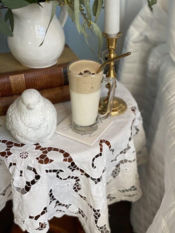 Picture of Dolgona coffee made with Javy coffee on and end table next to a chair. There is also some vintage books and a ceramic bird on the white table