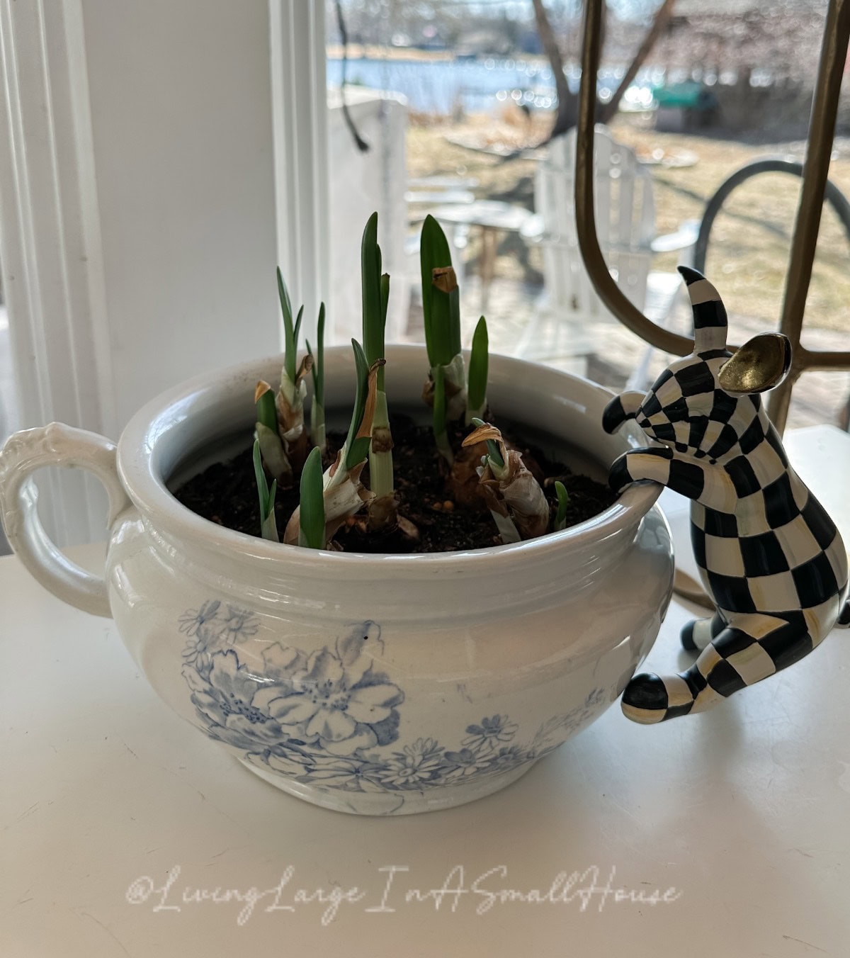 A blue and white ceramic pot with a floral design holds small green sprouts. A whimsical, budget-friendly black and white checkered cow figurine leans on the pot. The background reveals a window with a view of a garden, perfect for spring mantel decor.