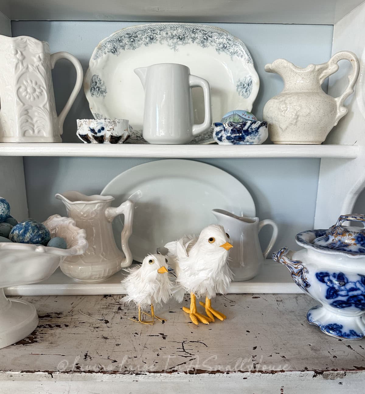 A whimsical collection of white and blue crockery graces the shelves, featuring plates, pitchers, and decorative bowls with floral patterns. Two fluffy, white bird ornaments with yellow beaks and feet stand on the worn wooden surface, offering budget-friendly spring mantel decor.