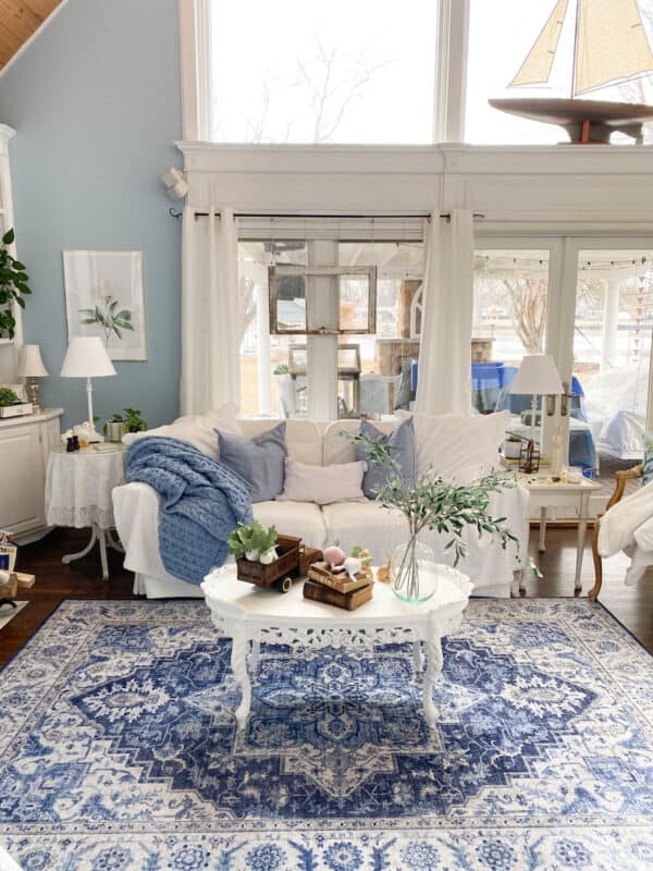 View of my living room with the white sofa against the window, a chair to the right and a white coffee table in front. There is a blue and white rug on the floor, blue and white pillows and a throw on the sofa. Moved all the furniture around as a refresh for spring