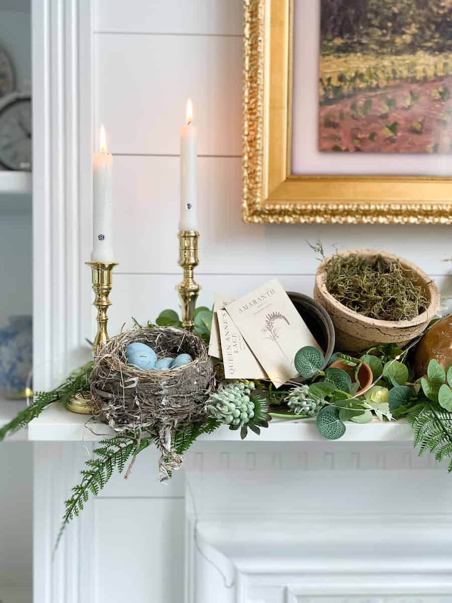 A decorated mantel for the Fabulous Friday Link Party 4.1.22 showcases two lit candles, a bird's nest with blue eggs, a botanical book, and a pot with moss amid lush green foliage. Above it all hangs a framed painting.